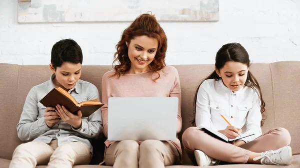 Femme souriante avec ordinateur portable assis près des enfants faisant leurs devoirs sur le canapé — Photo de stock