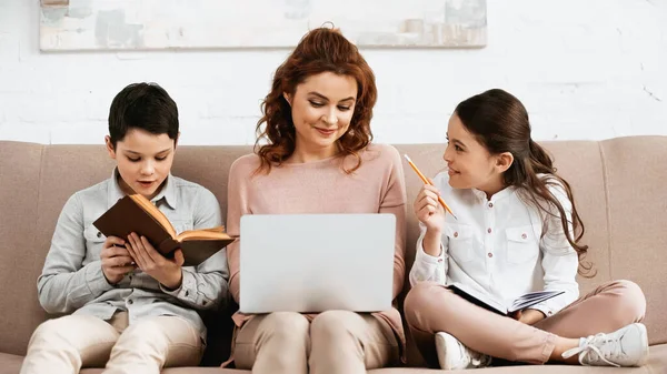 Веселая девушка с блокнотом и карандашом сидит рядом с матерью с ноутбуком и братом читает книгу — стоковое фото