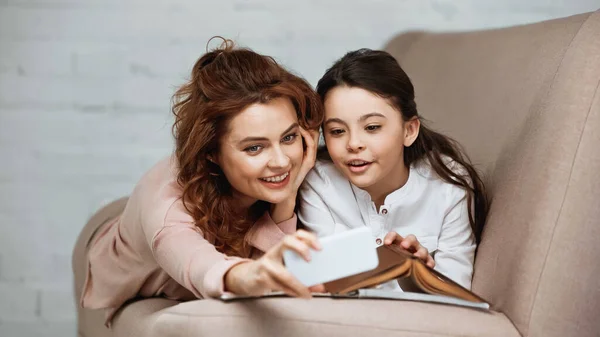 Sorrindo mãe tomando selfie com filha perto do livro em primeiro plano desfocado no sofá — Fotografia de Stock