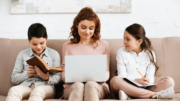 Smiling kids doing homework near mother with laptop on couch — Stock Photo