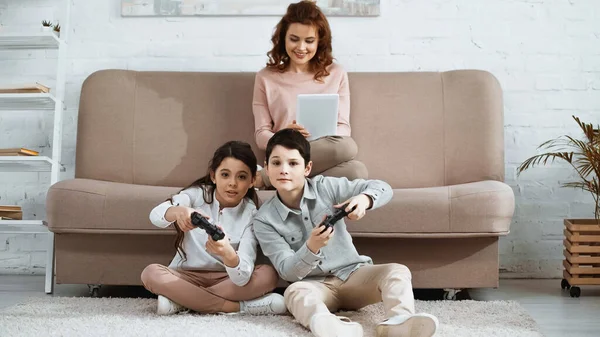 KYIV, UKRAINE - 15 AVRIL 2019 : Enfants jouant à un jeu vidéo près d'une mère souriante avec tablette numérique sur le canapé — Photo de stock