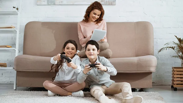 KYIV, UKRAINE - 15 AVRIL 2019 : Des enfants positifs avec des joysticks jouant à un jeu vidéo près d'une mère souriante avec une tablette numérique sur fond flou — Photo de stock