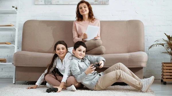 KYIV, UCRANIA - 15 DE ABRIL DE 2019: Familia alegre con joysticks y tabletas digitales mirando a la cámara en la sala de estar - foto de stock