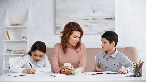 Mère avec smartphone assis près des enfants faisant leurs devoirs — Photo de stock