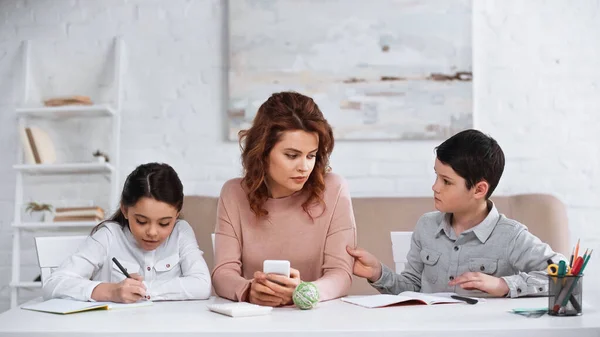Жінка з мобільним телефоном допомагає дітям з домашнім завданням за столом — стокове фото