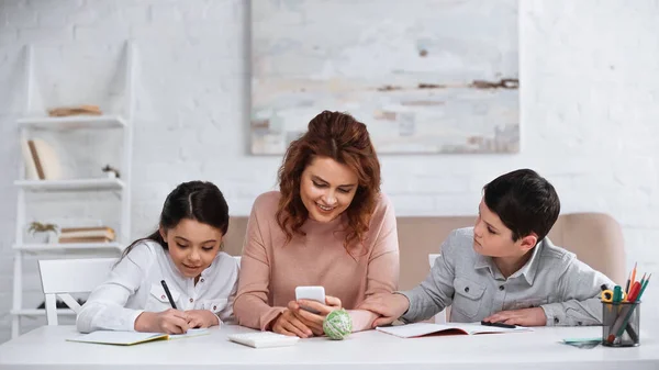 Lächelnde Mutter mit Smartphone bei Hausaufgaben mit Kindern im Wohnzimmer — Stockfoto