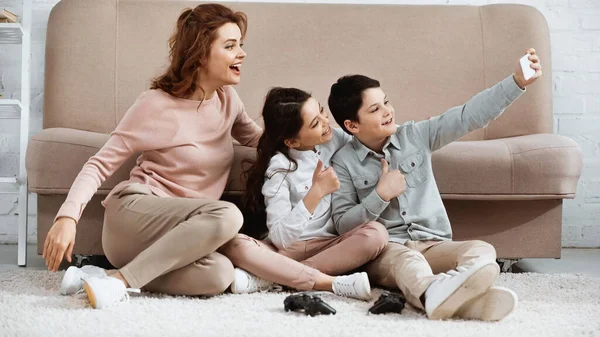 KYIV, UKRAINE -  APRIL 15, 2019: Smiling kids showing yes gesture while taking selfie near mother and joysticks on floor — Stock Photo