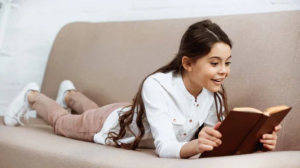 Positive kid reading book on couch — Stock Photo