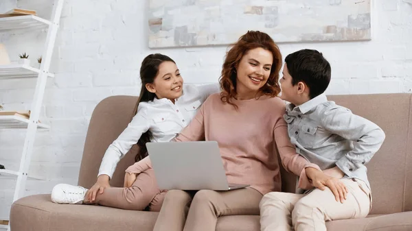 Sonriente madre con portátil abrazando a los niños en el sofá - foto de stock