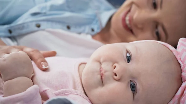 Baby girl looking at camera while lying near mother on blurred background — Stock Photo