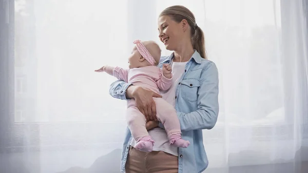 Freudige Mutter hält Baby-Tochter im Arm und zeigt mit der Hand auf Fenster — Stockfoto