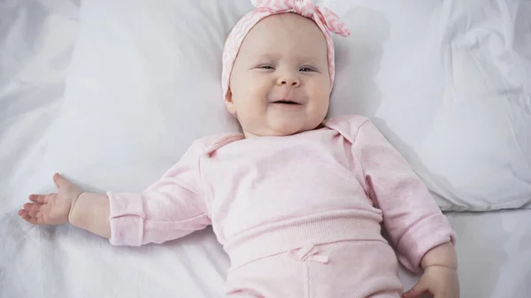 Top view of smiling infant girl in headband lying on white bedding — Stock Photo