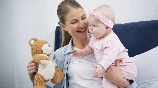Donna allegra che tiene il giocattolo morbido vicino alla figlia neonata in fascia con fiocco — Foto stock