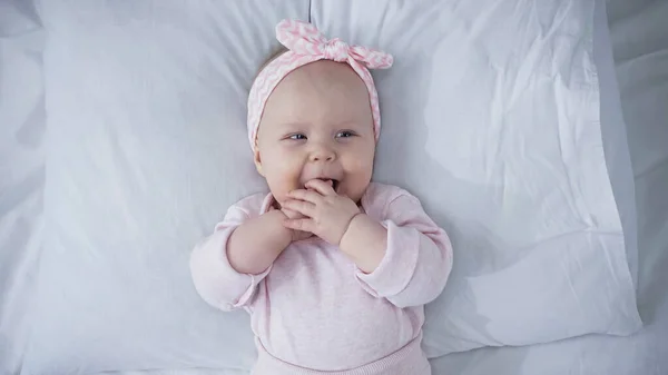 Top view of baby girl in headband with bow sucking fingers while lying on bed — Stock Photo