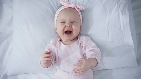 Top view of upset baby girl in headband with bow crying while lying on bed — Stock Photo