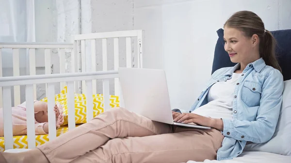 Happy mother using laptop near daughter in baby crib — Stock Photo