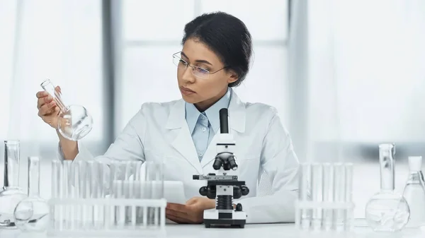 African american scientist in glasses holding flask near microscope — Stock Photo