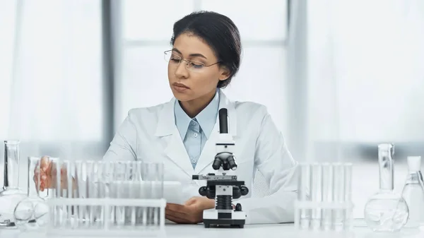 African american scientist in glasses taking flask near microscope in lab — Stock Photo