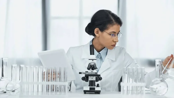Scientifique afro-américain dans des lunettes tenant tablette numérique près du microscope et des éprouvettes en laboratoire — Photo de stock