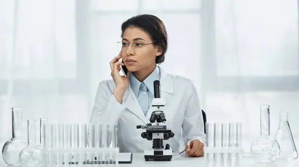 Афроамериканський вчений в окулярах розмовляє на смартфоні біля мікроскопа в лабораторії — стокове фото