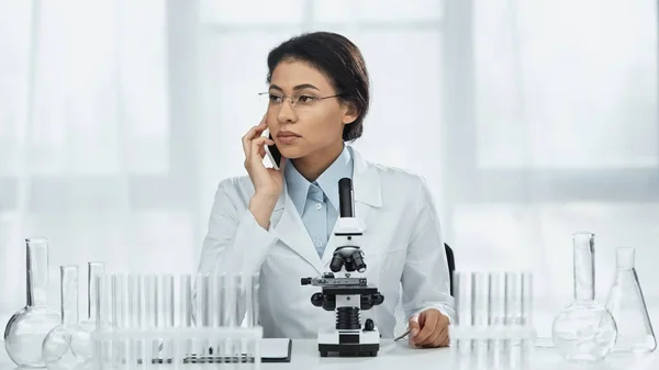 African american scientist in glasses talking on smartphone near microscope in lab — Stock Photo
