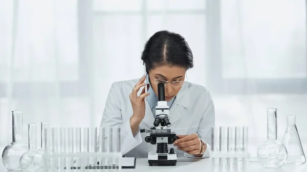Científico afroamericano en gafas hablando en smartphone mientras mira a través del microscopio en el laboratorio - foto de stock