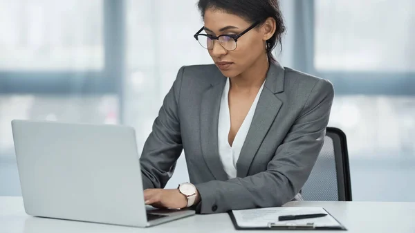 African american businesswoman in glasses using laptop near clipboard on desk — Stock Photo