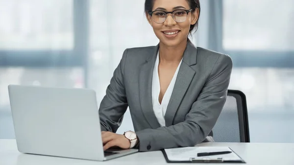 Feliz afroamericana mujer de negocios en gafas mirando a la cámara mientras se utiliza el ordenador portátil - foto de stock