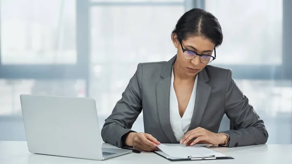 African american businesswoman in glasses looking at papers on clipboard near laptop — Stock Photo