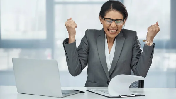 Emocionada mujer de negocios afroamericana en gafas cerca de la computadora portátil - foto de stock