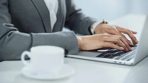 Vista cortada de empresária digitando no laptop perto da xícara de café em primeiro plano desfocado — Fotografia de Stock