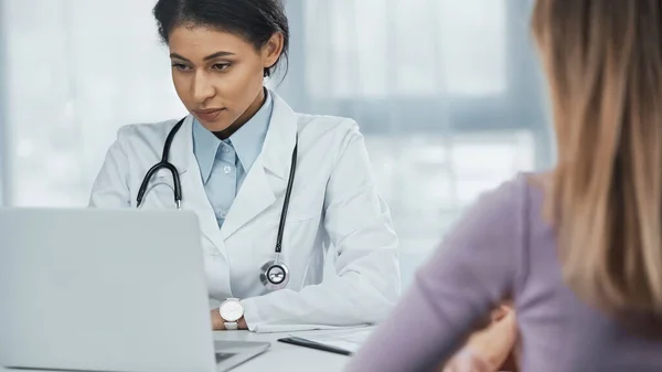 African american doctor in white coat using laptop near patient on blurred foreground — Stock Photo