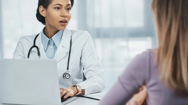 African american doctor in white coat using laptop and talking with patient on blurred foreground — Stock Photo