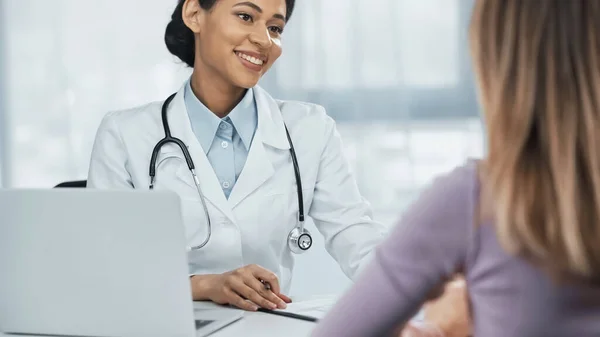 Счастливый африканский американский врач в белом халате разговаривает с размытым пациентом возле ноутбука на столе — стоковое фото