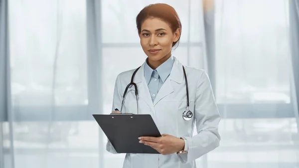 Cheerful african american doctor in white coat with stethoscope holding clipboard in clinic — Stock Photo