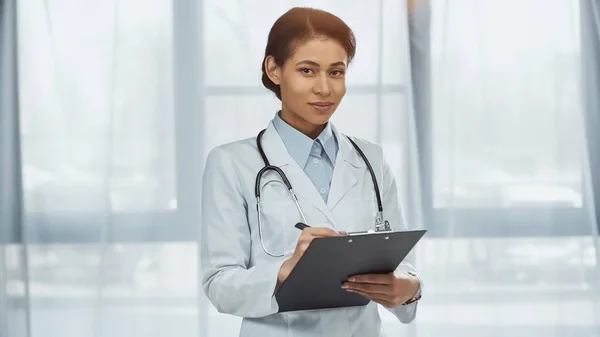 Happy african american doctor in white coat with stethoscope holding clipboard in clinic — Stock Photo
