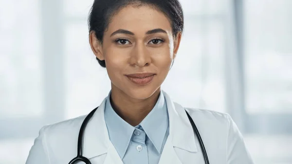 Fröhliche junge afrikanisch-amerikanische Ärztin im weißen Mantel mit Stethoskop blickt in die Kamera — Stockfoto