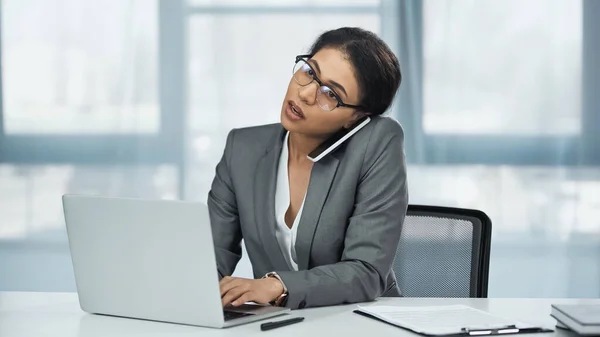 African american businesswoman in glasses talking on smartphone near laptop on desk — Stock Photo