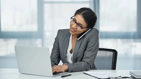 Feliz mujer de negocios afroamericana hablando en el teléfono inteligente cerca de la computadora portátil en el escritorio - foto de stock