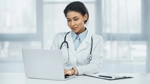 Smiling african american doctor using laptop near clipboard on desk — Stock Photo