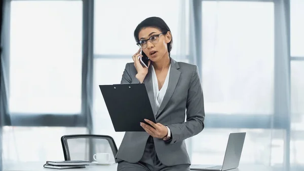 African american businesswoman talking on mobile phone and holding clipboard near laptop on desk — Stock Photo