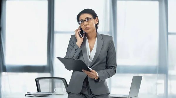 African american businesswoman in glasses talking on mobile phone and holding clipboard near laptop on desk — Stock Photo