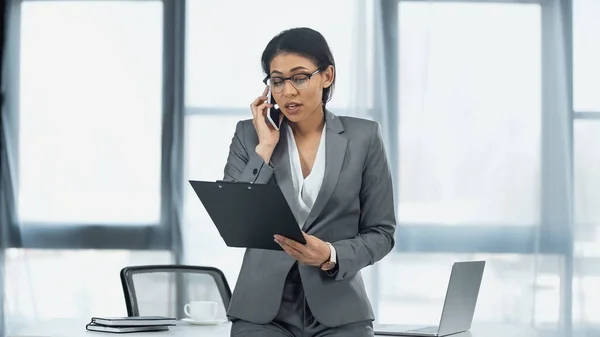 Mujer de negocios afroamericana hablando en el teléfono inteligente y sujetando portapapeles cerca de la computadora portátil en el escritorio - foto de stock