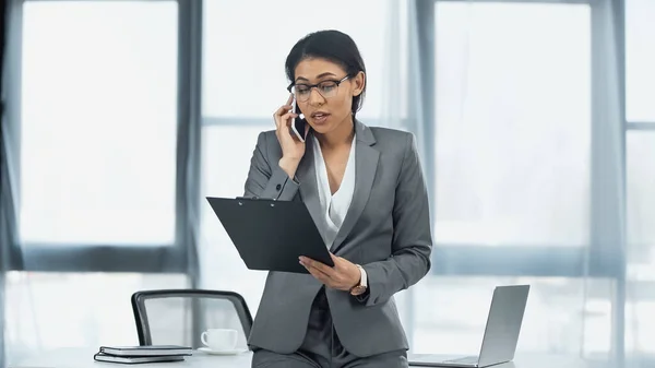 Afro-américaine femme d'affaires parlant sur smartphone et tenant presse-papiers près d'un ordinateur portable sur le bureau — Photo de stock