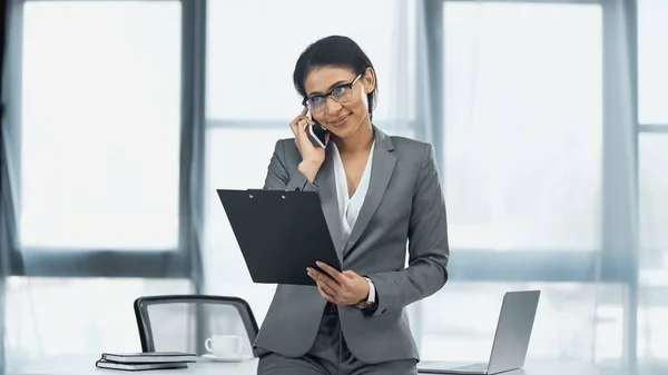 Mujer de negocios afroamericana feliz hablando en el teléfono móvil y sujetando el portapapeles cerca del ordenador portátil en el escritorio - foto de stock