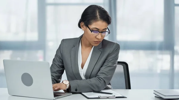 African american businesswoman typing on laptop while looking at clipboard — Stock Photo