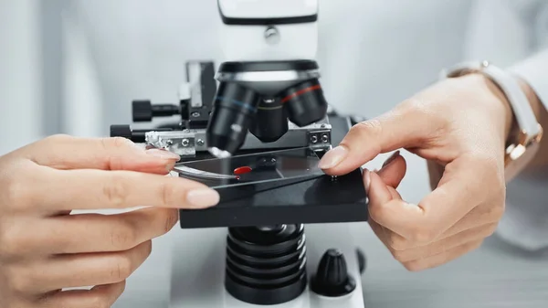 Abgeschnittene Ansicht eines afrikanisch-amerikanischen Wissenschaftlers, der im Labor eine Glasplatte mit Blutprobe in der Nähe des Mikroskops hält — Stockfoto