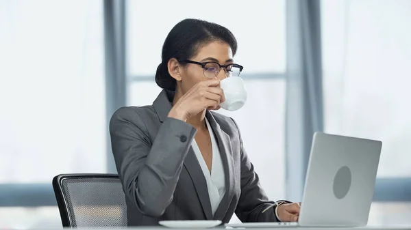 Afrikanisch-amerikanische Geschäftsfrau trinkt Kaffee in der Nähe von Laptop — Stockfoto