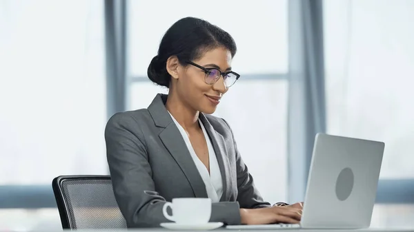 Feliz mulher de negócios afro-americana usando laptop perto de xícara de café — Fotografia de Stock