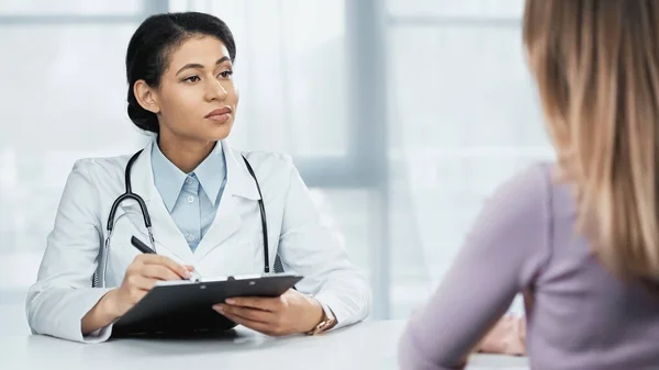 African american doctor in white coat writing diagnosis near patient on blurred foreground — Stock Photo